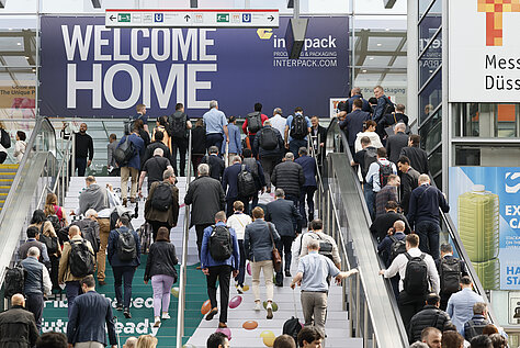 Una gran multitud de personas sube por las escaleras mecánicas hacia un cartel en el que se lee «BIENVENIDOS A CASA» en la entrada del evento Interpack.